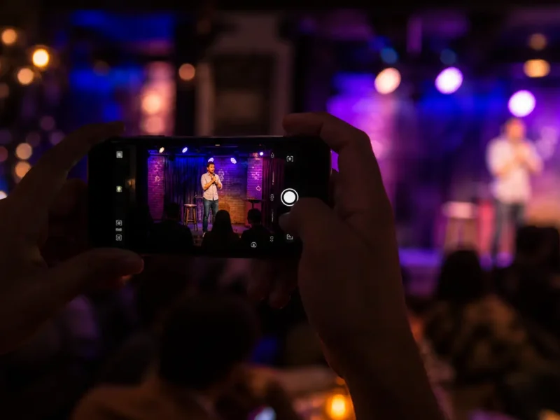 Person taking a photo of a live stand-up comedy show in Noida with a smartphone, dimly lit club stage and nightlife ambiance