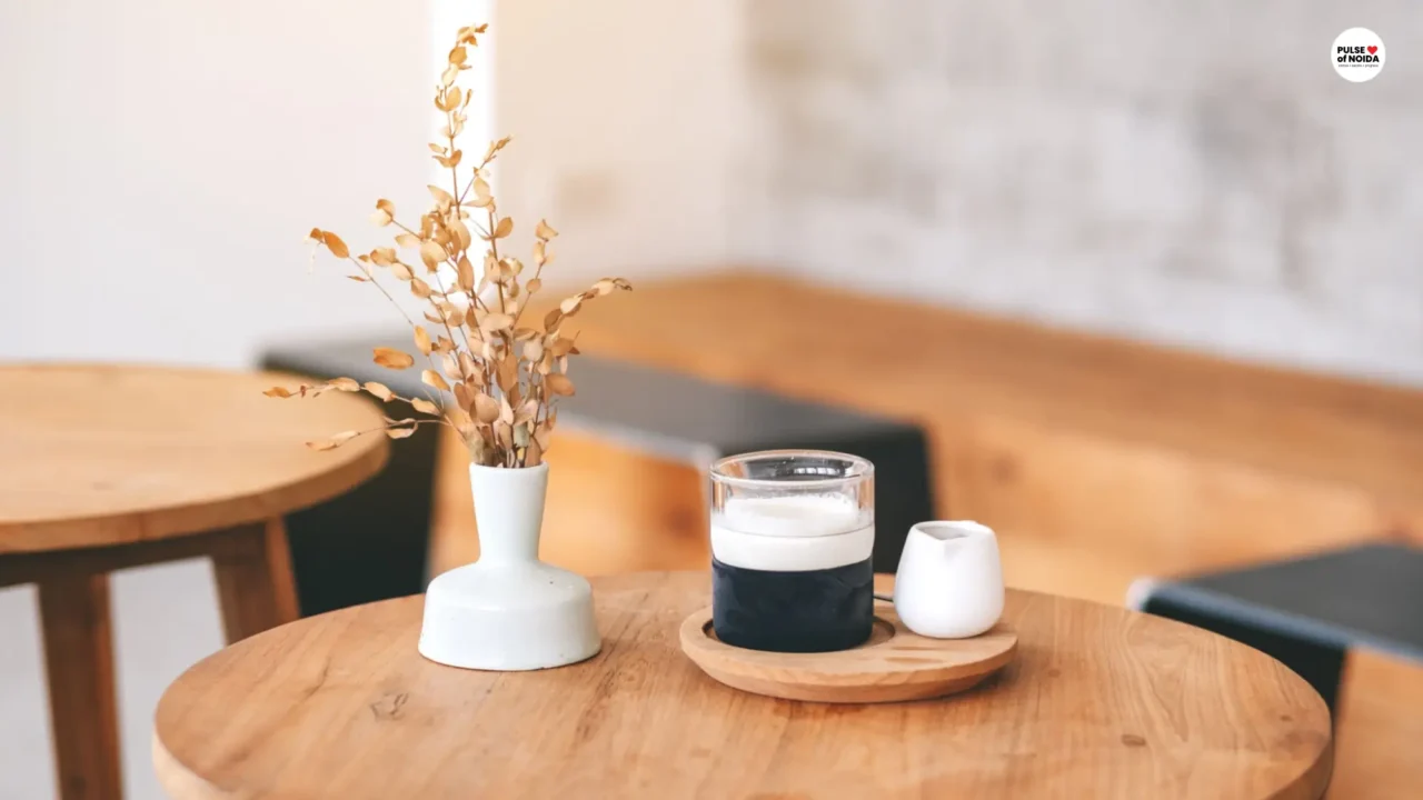 A glass of coffee and a white vase on a round wooden table inside a softly lit cafe.