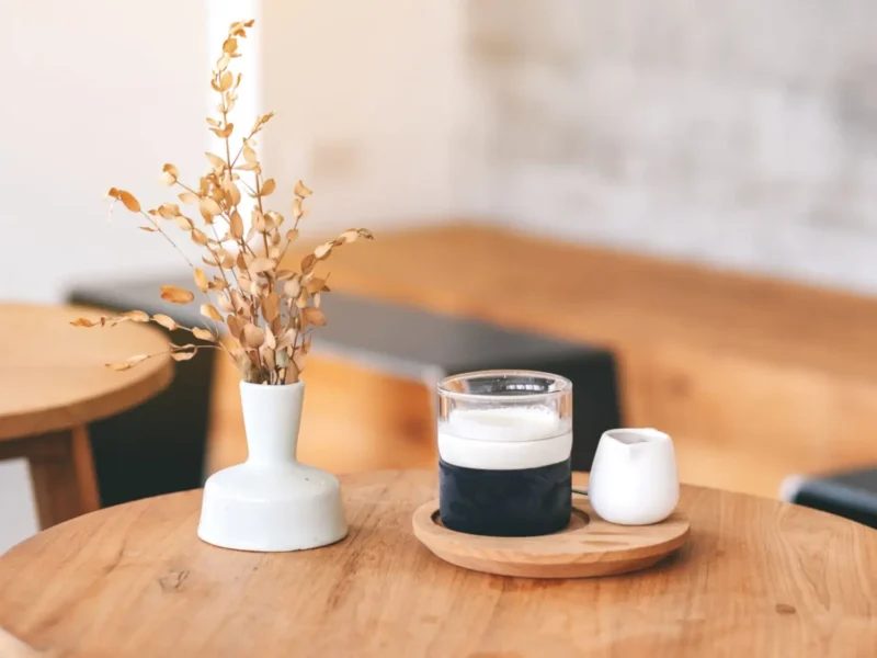 A glass of coffee and a white vase on a round wooden table inside a softly lit cafe.