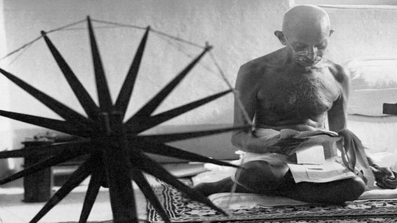 Mahatma Gandhi seated reading, with a spinning wheel in the foreground.