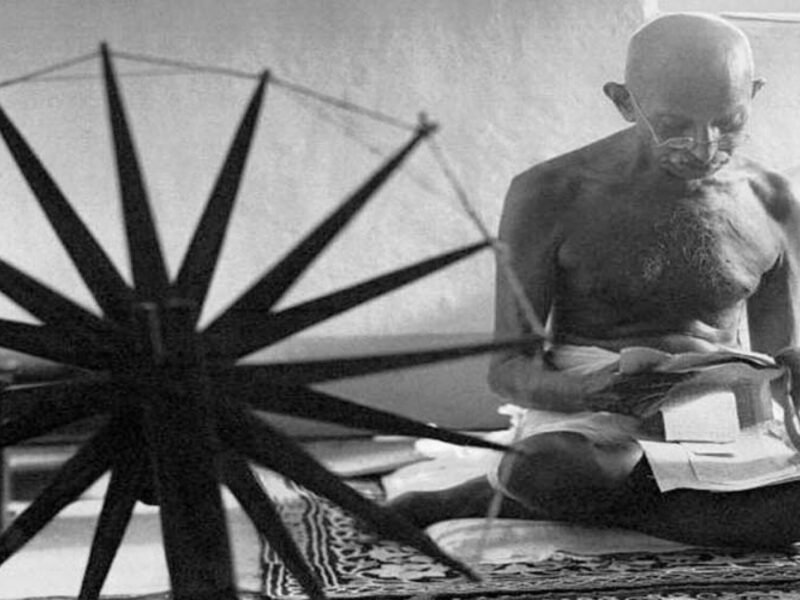 Mahatma Gandhi seated reading, with a spinning wheel in the foreground.